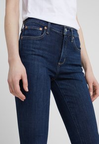 Pantalones vaqueros de mezclilla azul oscuro con un corte ajustado, featuring herrajes de cobre, cinco bolsillos y costuras en contraste. Textura suave y diseño clásico.