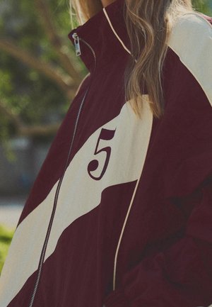 Persona con chaqueta con cremallera color granate y crema con el número 5 en el pecho, al aire libre con vegetación desenfocada en el fondo.