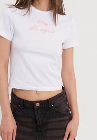 Hvid croppet t-shirt med pink broderet tekst "No regrets" og lille pink bånddetalje. Har bølget halsudskæring og korte ærmer.