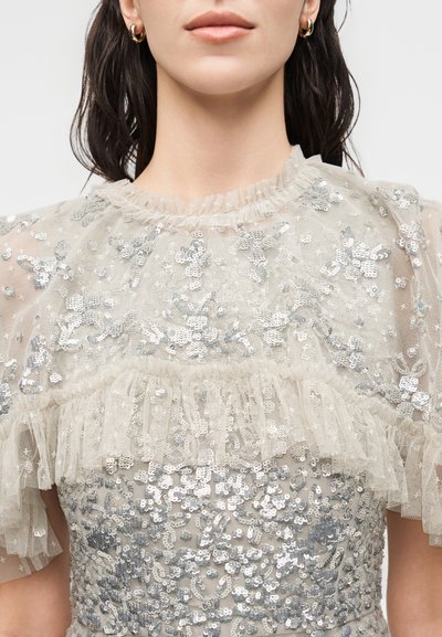 Γυναίκα που φοράει διάφανο μπεζ μπλουζάκι με ασημένια παγέτα λουλούδια και βολάν στο ντεκολτέ, συνδυασμένο με μικρά χρυσά στεφάνια σκουλαρίκια.