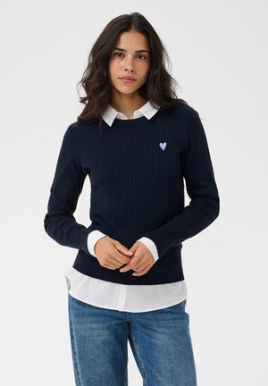 Femme aux cheveux foncés et ondulés portant un pull en maille torsadée bleu marine sur une chemise blanche à col et un jean bleu, debout, les mains jointes.