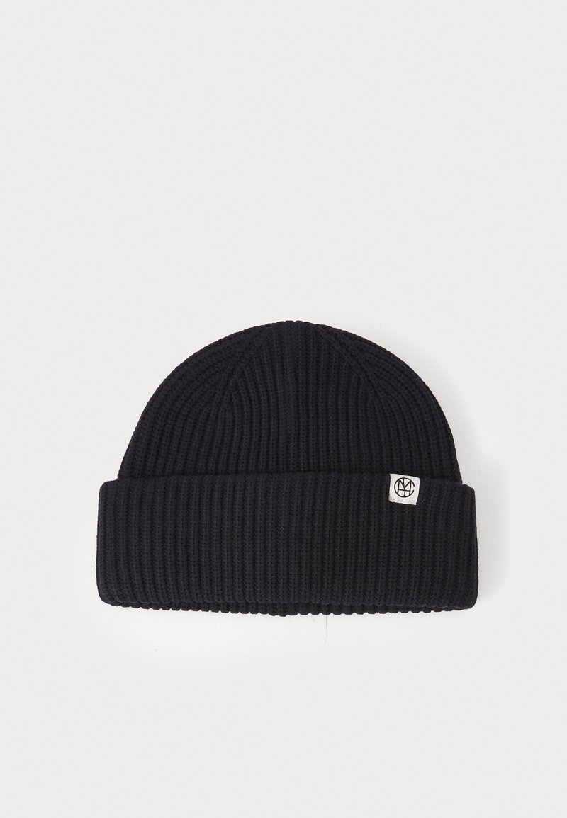 Bonnet tricoté bleu marine avec texture côtelée et revers. Présente un petit patch rond blanc sur le côté.