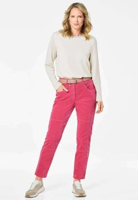 Roze corduroy broek met voorzakken en een bruine riem, gecombineerd met een lichtgekleurde lange mouwtop en grijze sportschoenen.