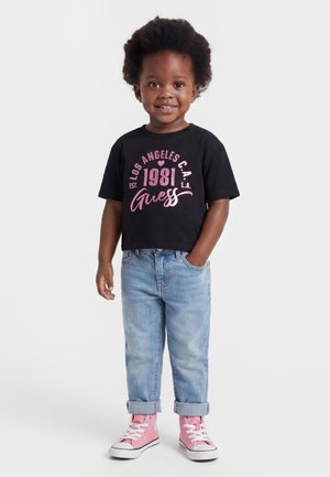 Småbarn med krøllet hår iført sort grafisk t-shirt, lyseblå optrukne jeans og lyserøde høje sneakers, smilende med den ene hånd i lommen.