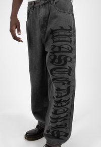 Mörkgrå denimbyxor med stora svarta gotiska bokstäver längs vänster ben. Löst snitt med en klassisk femfickdesign.