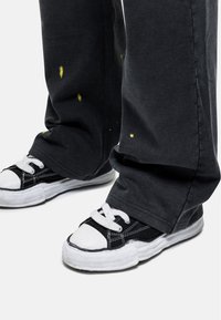 Zwarte broeken met verfspetterdetails, gecombineerd met zwart-witte laaggeschoeide sneakers met witte veters en rubberen zolen.