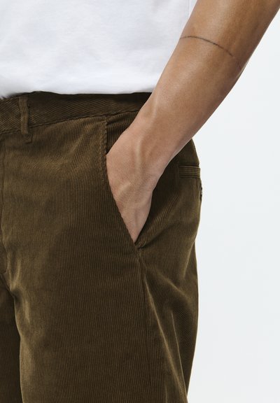 Pantalon en velours côtelé marron avec une texture à côtes verticales, poche latérale et taille ajustée. Le tissu semble doux et durable.