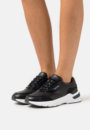 Baskets noires combinant du cuir et du daim, une semelle blanche épaisse, et des lacets noirs avec un logo sur la languette.