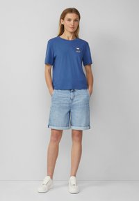 T-shirt à col rond bleu à manches courtes avec un petit motif sur la poitrine gauche, associé à un short en denim bleu clair retroussé au bas. Baskets blanches.