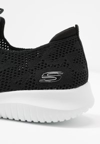 Mustat verkkomaiset urheilukengät, joissa on rakennepinta, valkoinen kumipohja ja metallinen logoaksentti sivulla. Kevyt malli, jossa on ilmanvaihto.