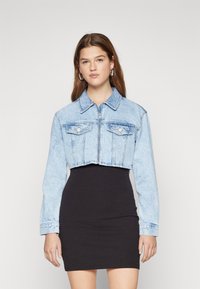 Lyseblå kort denimjakke med lange ermer, brystlommer og metallglidelås, sammen med en svart tettsittende kjole.