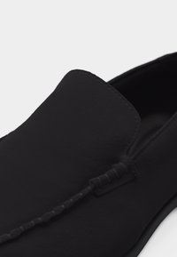 Chaussure noire à enfiler avec un bout arrondi et un détail de couture visible le long du bord supérieur et de la couture centrale.