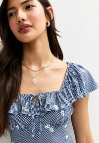 Blå rutig topp med volangärmar, blommiga detaljer och knut framtill. Guldlayerade halsband och slätt hår fulländar looken.