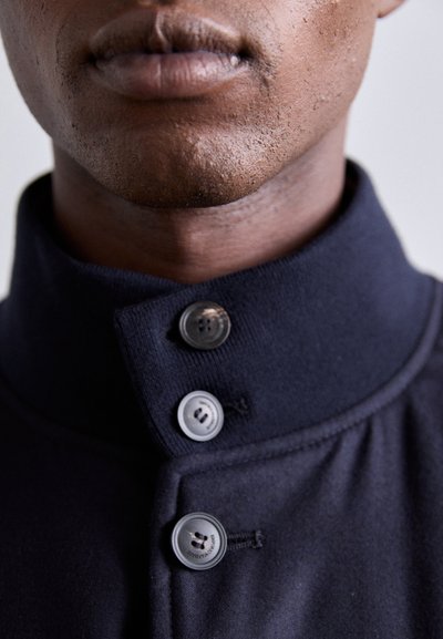 Marinblå fleeceplagg med hög krage och tre svarta knappar. Tyget har en slät yta och en ren, minimalistisk design.
