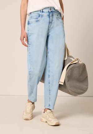 Persona che indossa jeans larghi azzurri chiari, sneakers bianche chunky e tiene una grande borsa da viaggio grigia con tracolle crema.