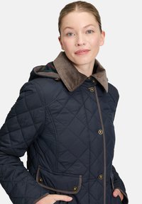 Marineblå quiltet jakke med en brun cordfløjskrave, gyldent hardware og to forlommer. Foret hætte har et kontrasterende ternet mønster.