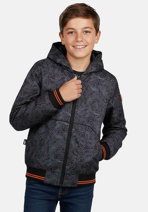 Lachende jongen die een zwarte capuchonjas draagt met een grijs topografisch patroon en oranje gestreepte manchetten, poserend met één hand bij de rits.