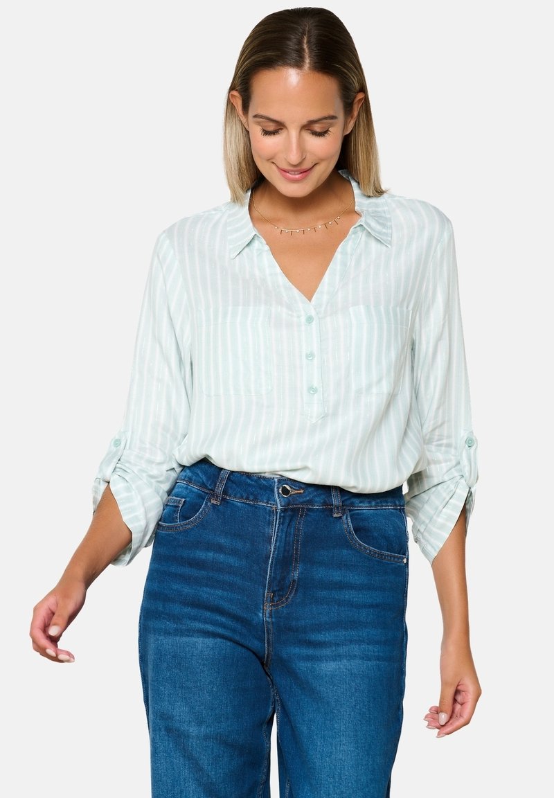 Camicia botton-down verde menta chiaro con strisce bianche verticali, tasche sul petto e maniche arrotolate, abbinata a jeans blu a vita alta.
