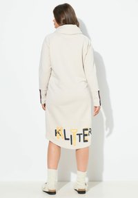 Beige, lange mouw sweatshirt jurk met een hoge kraag, voorzien van een "KLITTER" grafiek in zwart en geel aan de onderkant, gedragen met gebreide beenwarmers.
