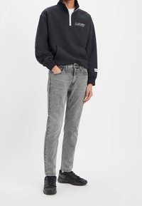 Sudadera negra con cremallera y logo blanco de Levi's, combinada con jeans grises de corte clásico y zapatillas deportivas negras. Diseño minimalista.
