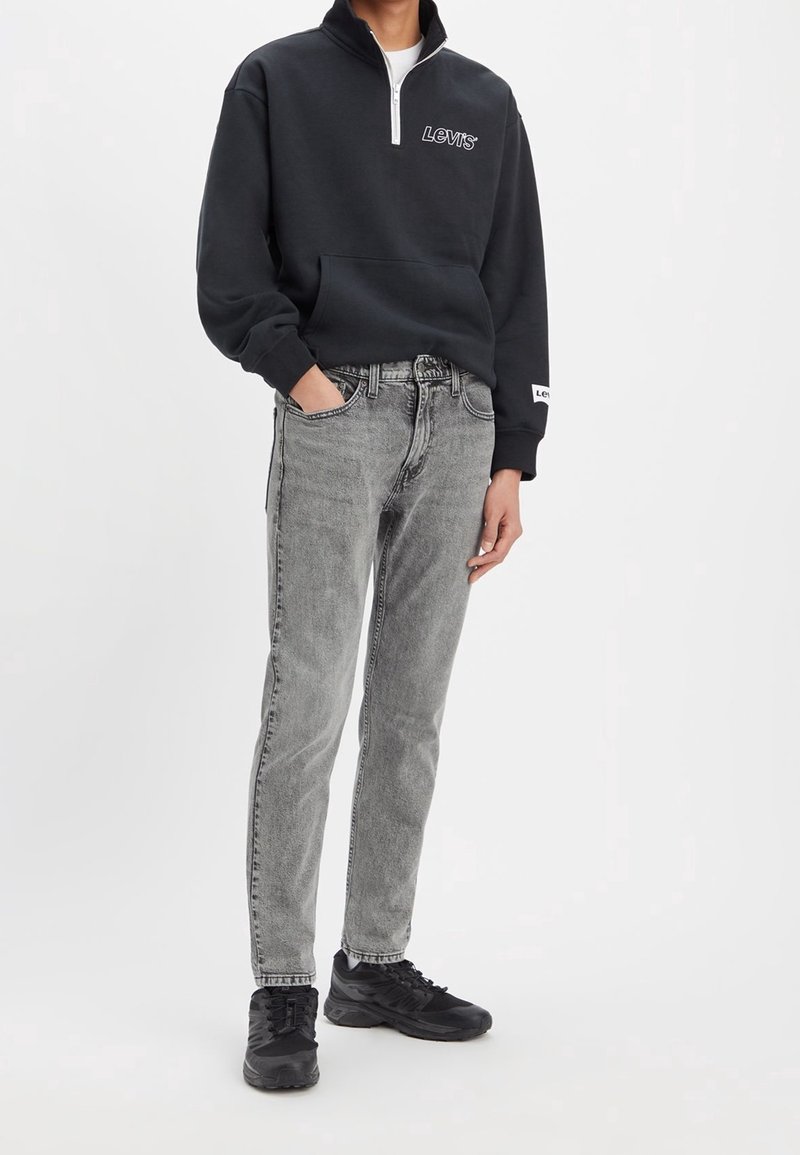 Sudadera negra con cremallera y logo blanco de Levi's, combinada con jeans grises de corte clásico y zapatillas deportivas negras. Diseño minimalista.