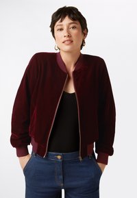 Giacca bomber in velluto bordeaux con polsini e colletto a costine, chiusura con zip, indossata sopra una maglia nera e abbinata a jeans blu scuro.