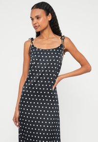 Melna satīna kleita ar baltām punktotu. Ir regulējamas lencītes ar saistāmiem akcentiem. Gluda tekstūra un plūstošs dizains.