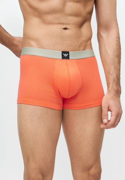 Männlicher Oberkörper, der eng anliegende orangefarbene Boxershorts mit hellgrauem Bund und schwarzem Logopatch in der Mitte vorne trägt.
