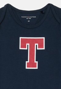Sötétkék baba rugdalózó, elöl nagy piros és fehér T betűvel, Tommy Hilfiger márkájú, 68-as méretben és alapítási dátuma: 1985.