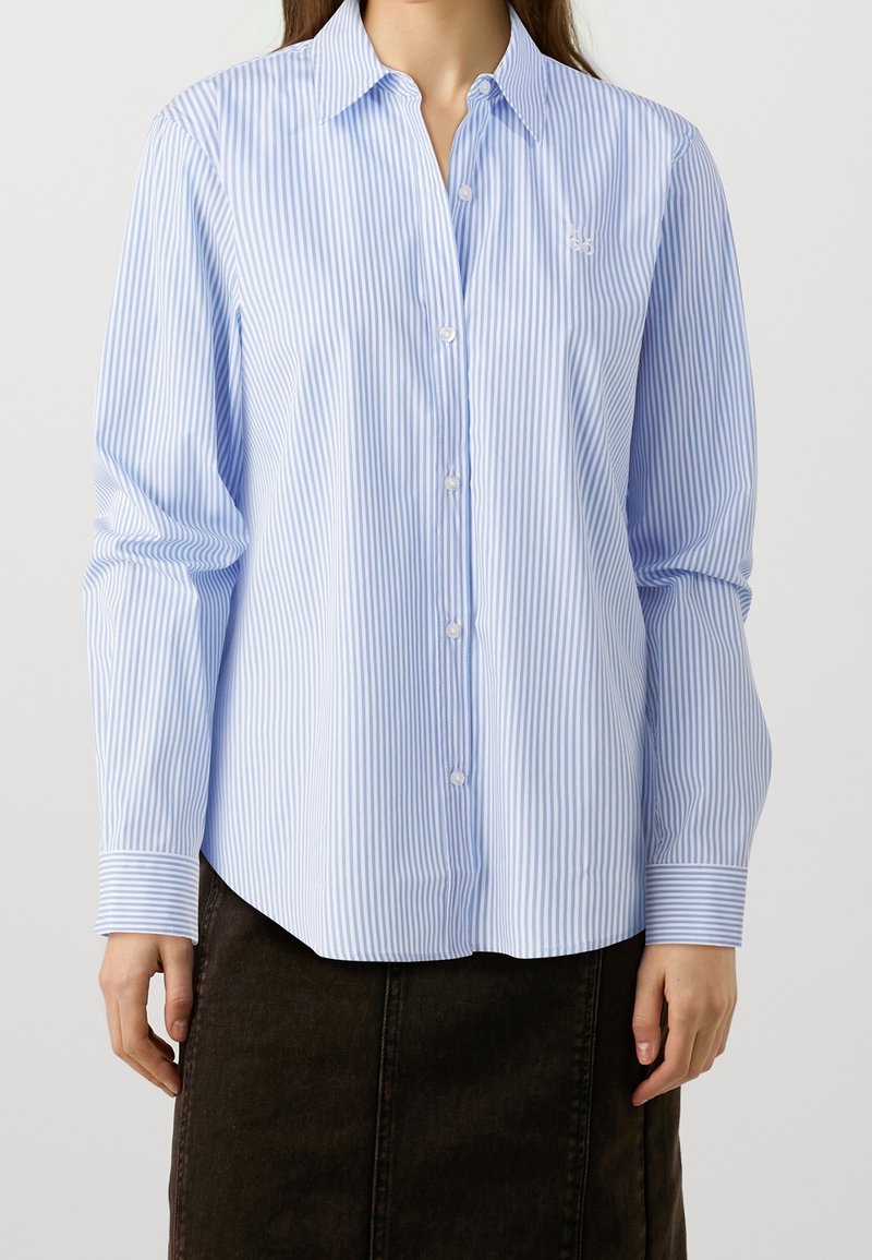 Femme portant une chemise à rayures verticales bleu et blanc avec boutons et manches longues, associée à une jupe foncée sur un fond uni.