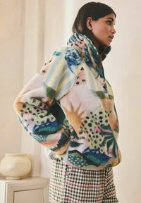 Femeie purtând o jachetă de fleece cu model colorat și pantaloni cu carouri verzi, stând lateral cu mâinile în buzunarele jachetei, în interior.