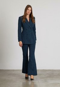 Blazer azul marino de doble botonadura con tejido texturizado, combinado con pantalones acampanados a juego, ambos con patrones intrincados. Incluye tacones altos negros.
