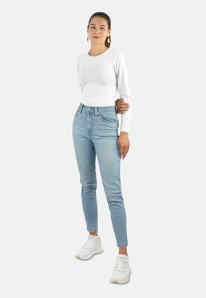 Junge Frau mit braunem Haar im Pferdeschwanz trägt ein weißes Langarmshirt, hellblaue hochtaillierte Jeans und weiße Sneakers und steht vor einem weißen Hintergrund.