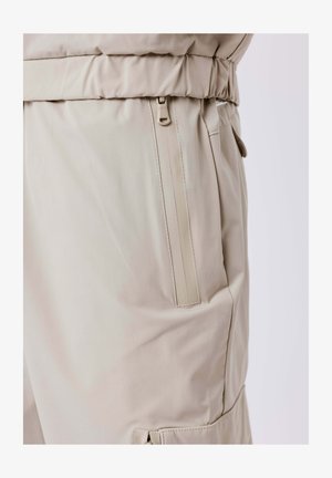 Pantalon beige clair avec une poche zippée sur le côté, une taille élastique et une texture de tissu lisse.