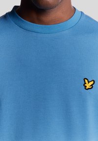 Uomo che indossa una maglietta blu a girocollo con un piccolo logo di un uccellino giallo sul lato sinistro del petto.