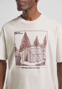 T-shirt beige chiaro con una grafica marrone di una tenda e alberi, con la scritta "Making room to roam" sotto, realizzata in morbido cotone.