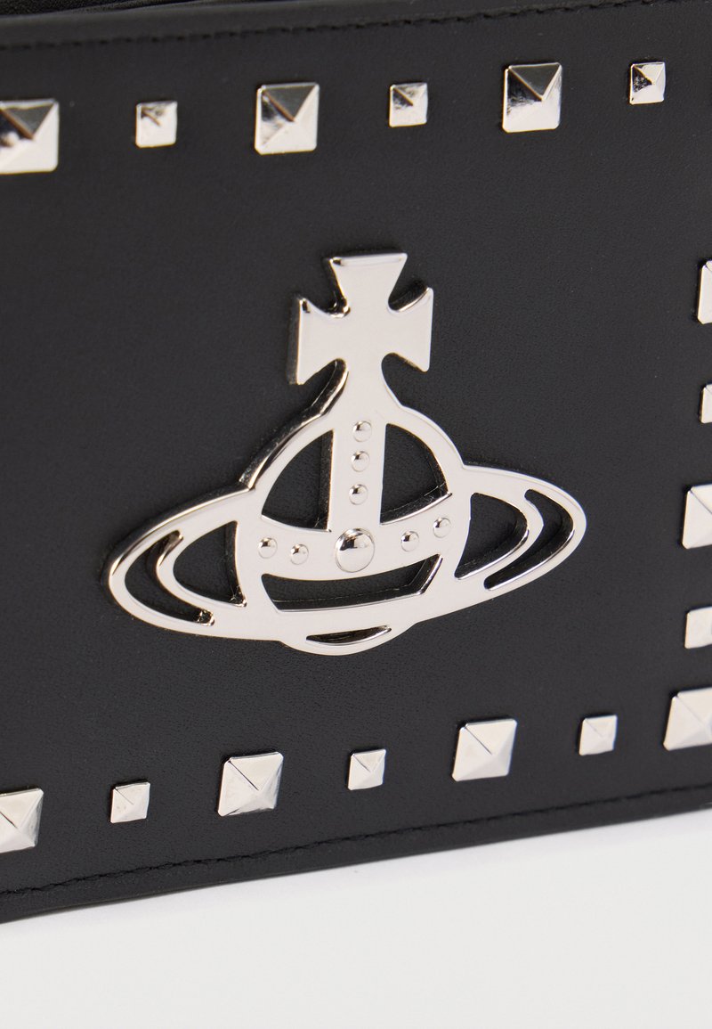 Surface en cuir noir avec un logo orbe et croix argentés, entouré de petits clous métalliques carrés disposés en rectangle.
