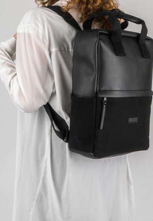 Sac à dos noir au design structuré, fabriqué en cuir et toile. Dispose de deux poignées supérieures, de bretelles ajustables et d'une poche zippée sur le côté.