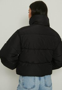 Melna īsā pūkainā jakas ar augstu apkakli, ar izšūtu dizainu un biezu izolāciju. Nēsāta pār gaiši ziliem džinsiem.