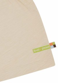 Camicia di cotone beige chiaro con orlo arrotondato e etichetta verde "loud + proud" con accenti colorati sul lato. Texture morbida.