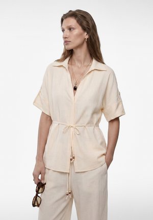 Vrouw met lang bruin haar draagt een crèmekleurig kortärmelig shirt en broek, houdt tortoisezonnebril in één hand en kijkt naar links.