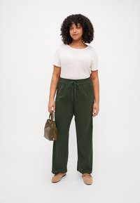 Camiseta corta de manga corta en beige claro, pantalones verdes con cintura ajustable y zapatos beige. Sosteniendo un pequeño bolso de mano en verde oliva. Diseño sencillo, ajuste relajado.