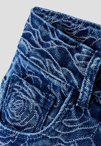 Denim tkanina s izvezenim cvjetnim uzorcima u bijelom koncu, s prikazom džepa od traperica i detaljima petlje za pojas.