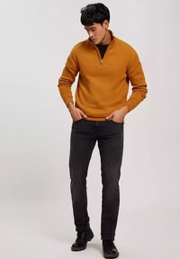 Musztardowy sweter z zamkiem ćwiartkowym, ze spiętymi mankietami i dołem, noszony z czarnymi dżinsami i czarnymi butami. Materiał wygląda na miękki z teksturowanym wykończeniem.