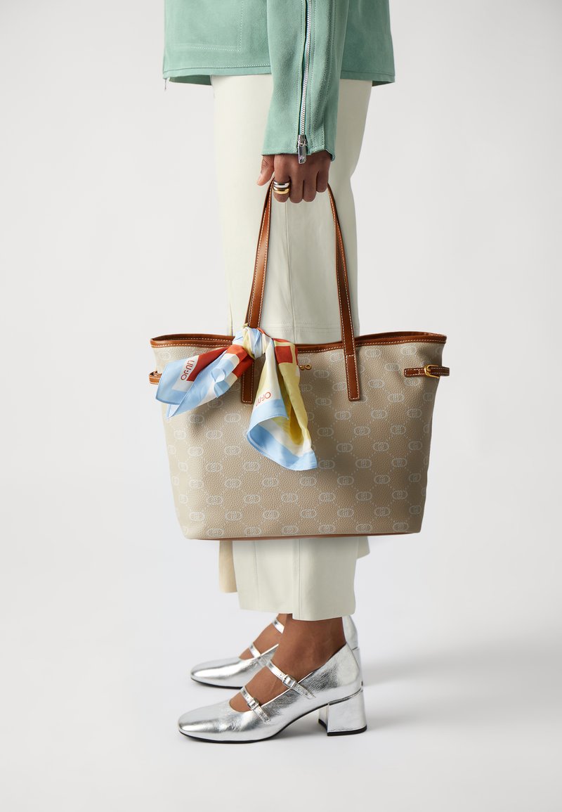 Borsa a tracolla beige con motivo e manici marroni, con una sciarpa blu a motivo annodata. Il modello indossa pantaloni chiari e décolleté Mary Jane argentati.