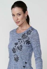 Kék, textúrált, hosszú ujjú felső, amelynek bal oldalán fekete virágmotívum hímzés található, az alsó jobb oldalon pedig gombos díszítések.
