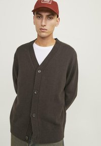 Cardigan a maglia di colore marrone scuro con scollatura a V, polsini a coste e cinque bottoni sul davanti. Indossato sopra una t-shirt bianca.