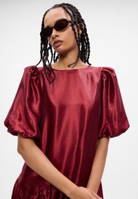 Βουργουνδί σατέν φόρεμα με φουσκωτά μανίκια και βολάν στο τελείωμα. Το μοντέλο φοράει σκούρα γυαλιά ηλίου. Λείος υφασμάτινος ιστό με γυαλιστερό φινίρισμα.