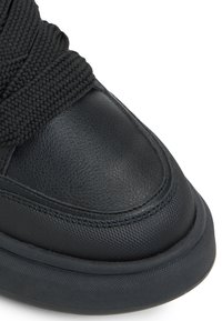 Detalle de zapatilla deportiva negra de cuero con parte superior texturizada, puntera lisa, cordones de tela trenzada y suela de goma redondeada.