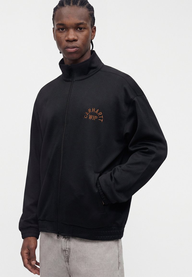 Junger Mann mit geflochtenem Haar trägt eine schwarze Zip-up-Jacke mit orangefarbenem „Carhartt WIP“-Logo, die Hände in den Taschen, kombiniert mit hellgrauen Hosen.